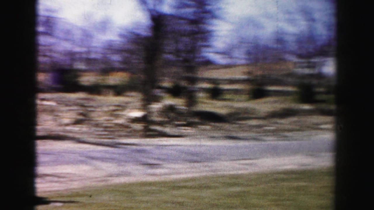 Continued 8mm pan across a 1950s suburban neighborhood with a road, sparse lawn, bare deciduous trees, residential houses, and a possible period automobile.