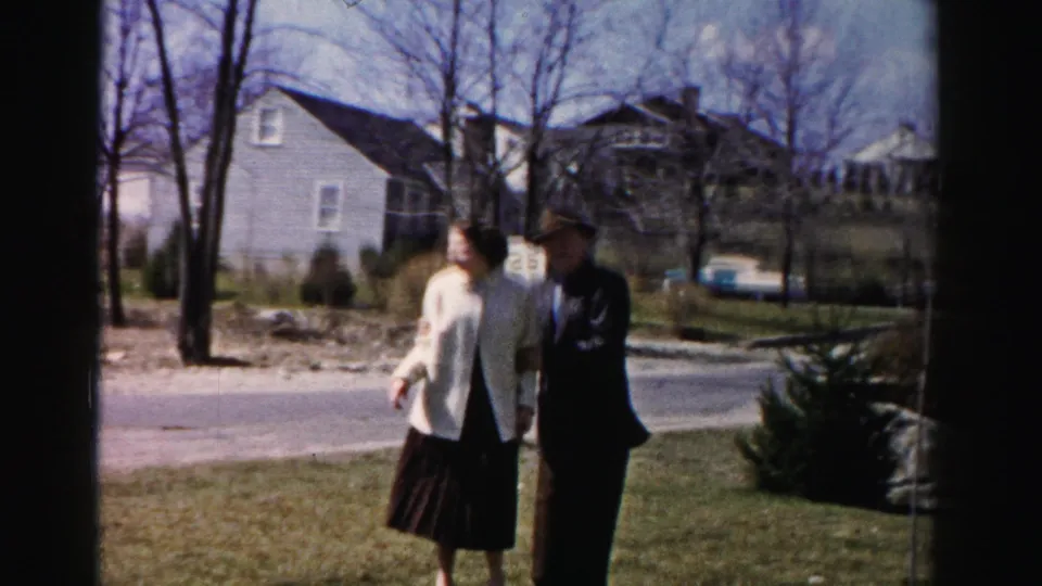 A couple on a suburban lawn in mid-1950s Albany, New York — woman in white boxy jacket and dark skirt, man in dark suit — with bare trees, a grey house, and a teal period automobile behind them.