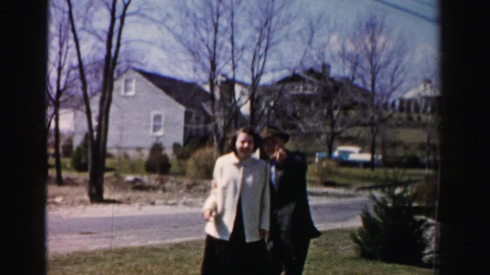 The same couple walks toward the camera on a suburban lawn in mid-1950s Albany, New York; a light-colored period automobile and grey house are visible in the background.