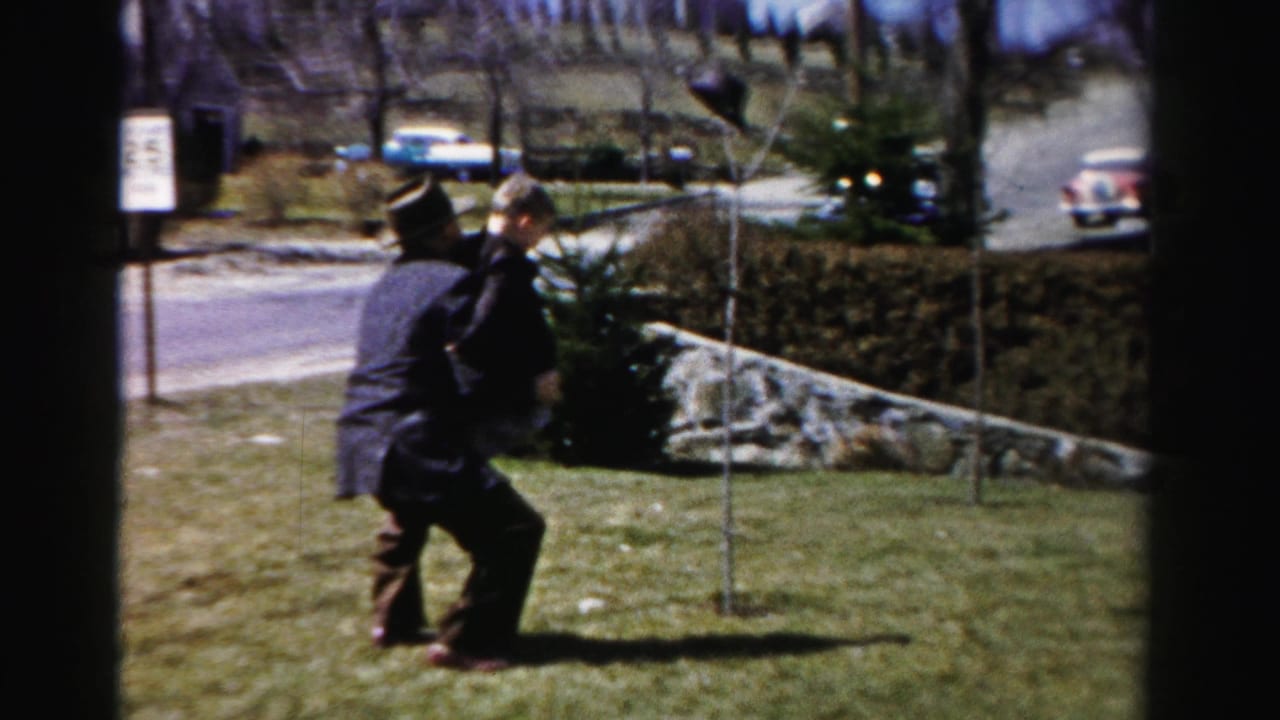 Man in 1950s dark suit and fedora-style hat lifts a small child near a stone-edged hedgerow on what appears to be a residential Albany, New York street.