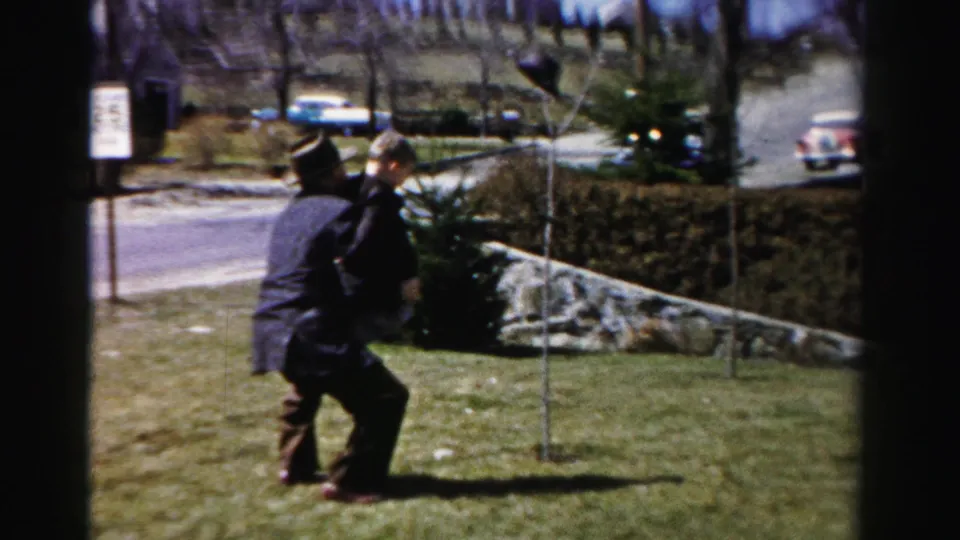 Man in 1950s dark suit and fedora-style hat lifts a small child near a stone-edged hedgerow on what appears to be a residential Albany, New York street.
