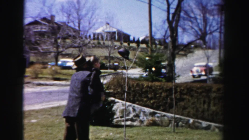 Man in 1950s attire and small child on a residential lawn in Albany, New York; stone-edged hedge, period automobiles, and bare trees fill the background.