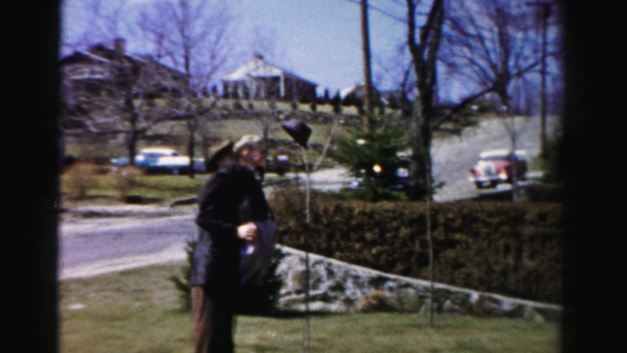 Man in dark jacket and brimmed hat on a suburban Albany, New York lawn, circa mid-1950s, with period automobiles visible on the residential street behind a manicured hedge.