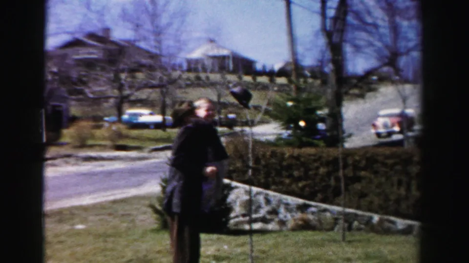 Man in dark jacket and hat with a second figure on a suburban Albany, New York lawn, circa mid-1950s; residential street with period automobiles visible behind a hedge.