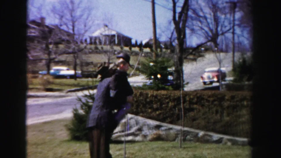 Man in dark jacket and brimmed hat appears to carry a small figure on a suburban Albany, New York lawn, circa mid-1950s; classic American automobiles parked on the residential street behind a hedge.