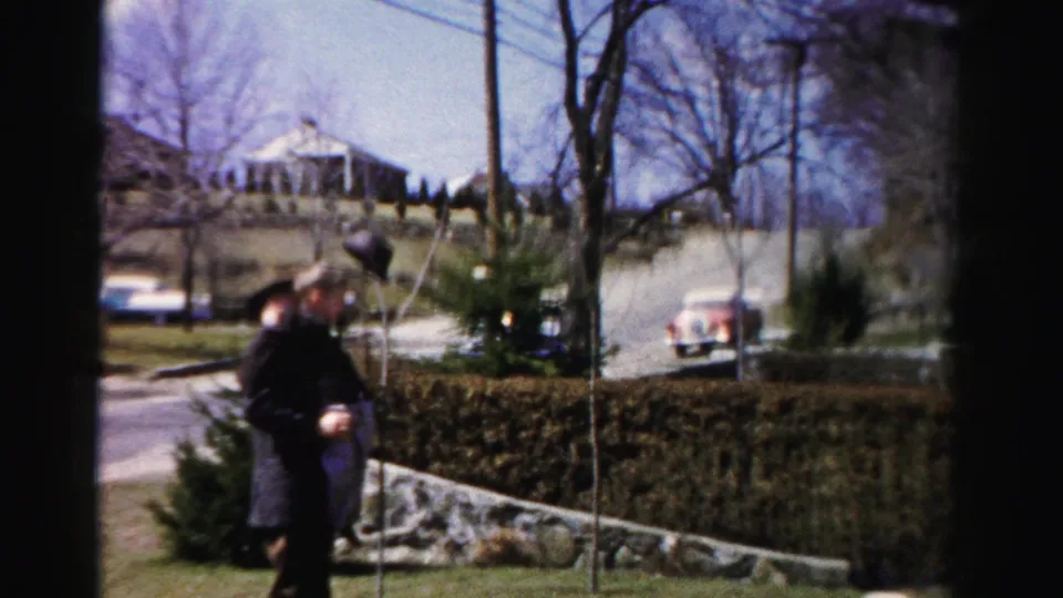 Adult male in dark overcoat bending toward a young child in a suburban yard, Albany, New York, circa 1955-1956; red mid-century automobile visible beyond the hedge row.