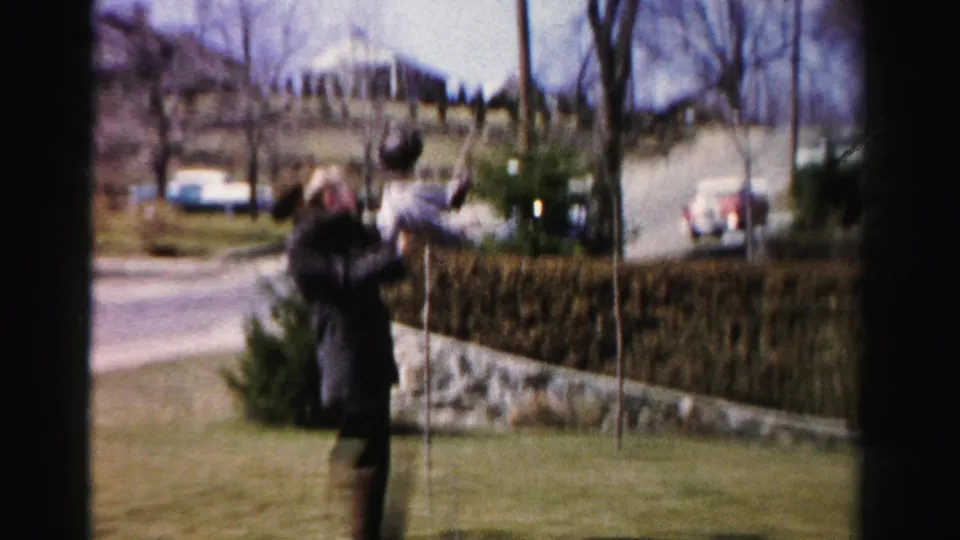 Adult male in dark overcoat lifting a young child in a playful gesture in a suburban yard, Albany, New York, circa 1955-1956; mid-century automobiles and residential street visible in background.
