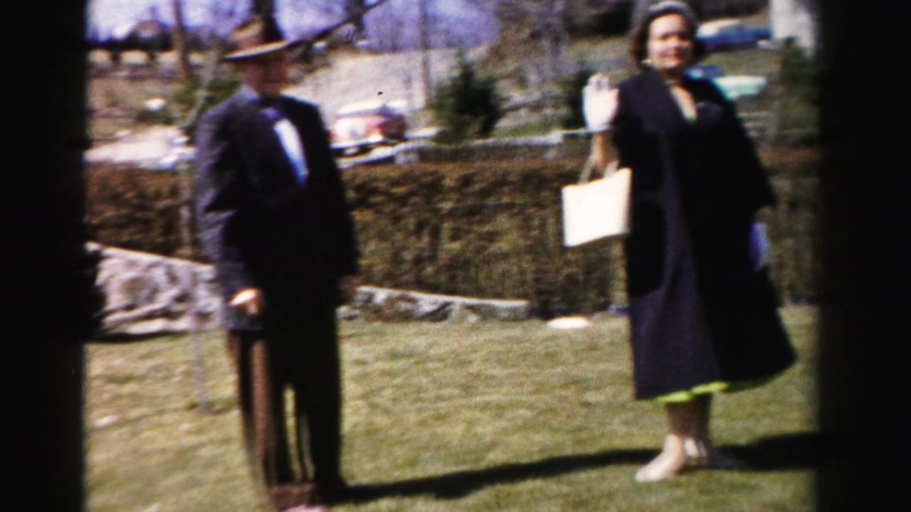 Mid-1950s color 8mm frame: man in fedora and dark suit stands beside woman in dark navy coat carrying a light box purse, waving at camera; grassy suburban lawn with stone border and hedgerow behind.