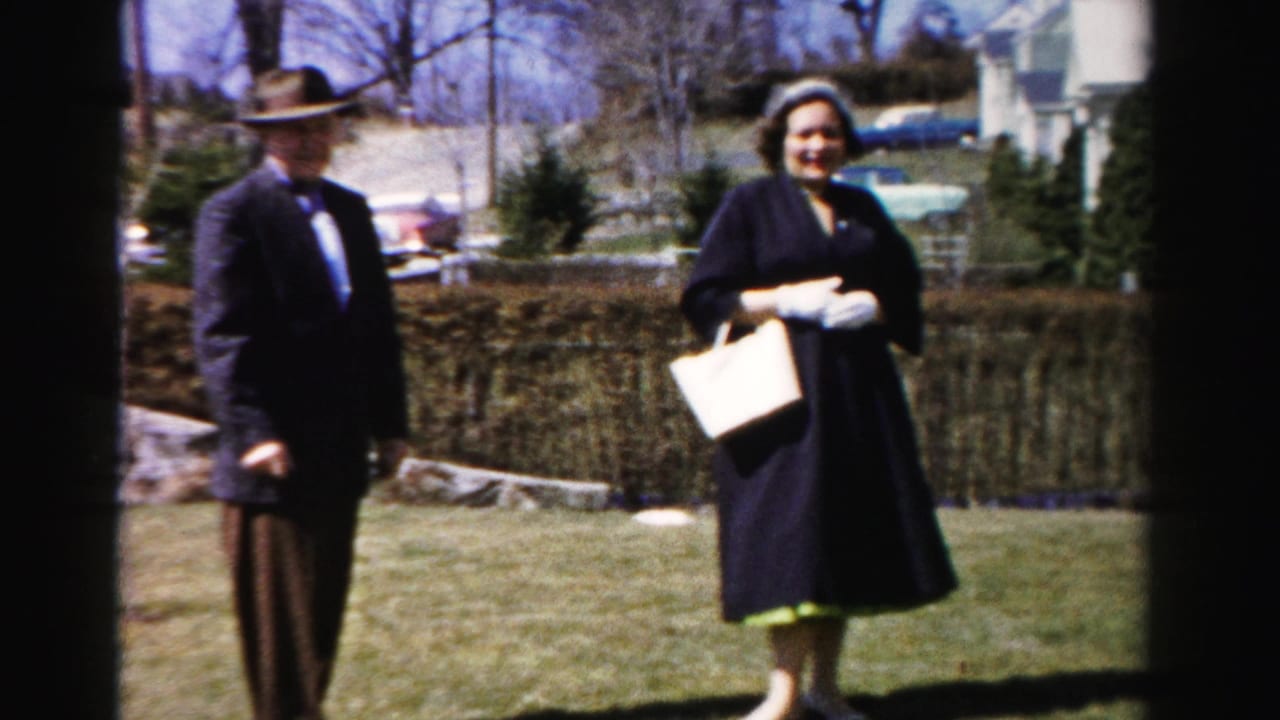 Man in dark suit and hat stands beside smiling woman in dark coat holding purse on sunny suburban lawn; partial red car visible in background.