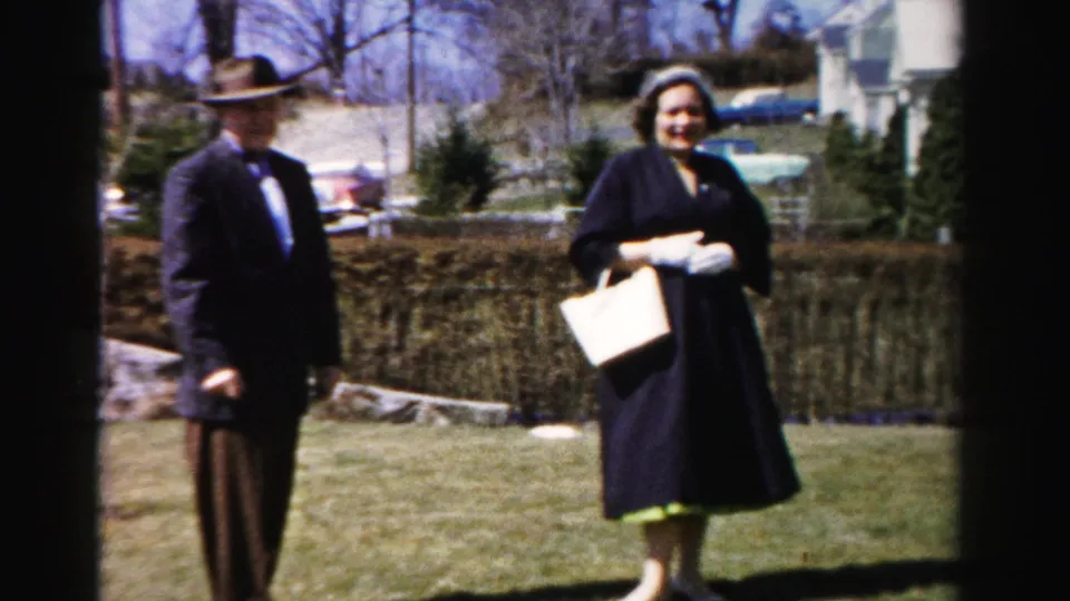 Color 8mm home movie: man in wide-brim fedora and dark suit alongside woman in dark coat with light purse smiling toward camera; stone border, bare trees, and partial automobile visible behind them.