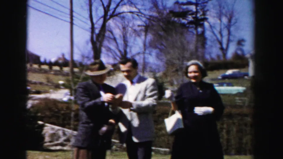 Color 8mm frame showing three adults on a suburban lawn: older man in dark suit and hat, younger man in light gray jacket, and woman in dark coat; bare trees, utility pole, and distant outbuilding visible.