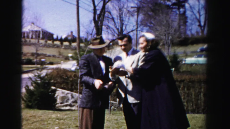 Color 8mm home movie frame: older man in fedora and dark suit, younger man in light jacket, and woman in dark coat in close animated interaction on a suburban lawn; bare trees and blue sky in background.