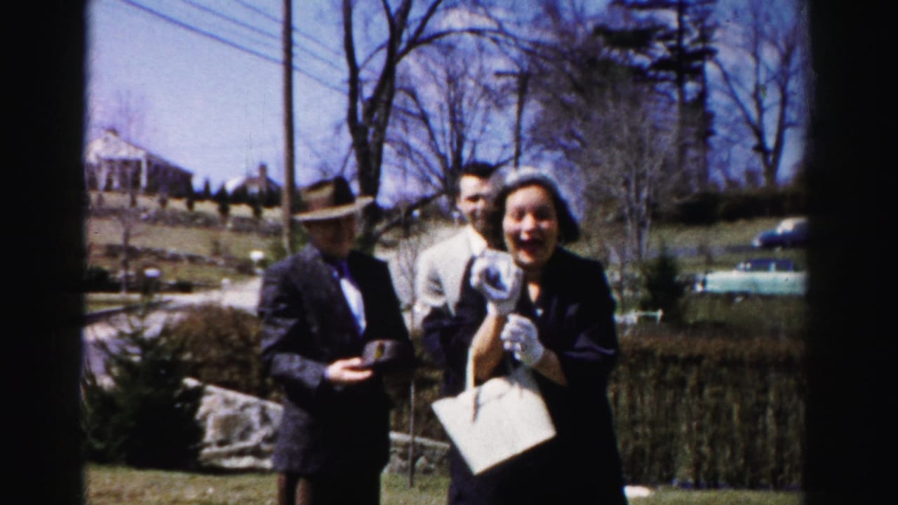 Woman in a dark coat laughs broadly outdoors; man in a dark fedora stands beside her, white structured handbag visible at her side.
