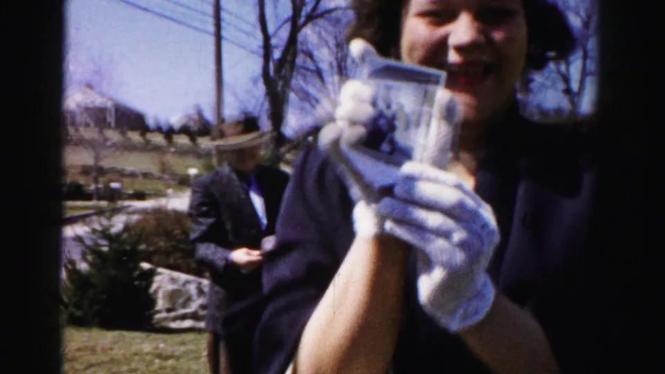 Close-up of a woman wearing white cotton gloves extending a small photograph or card toward the camera with a smile; a man in dark clothing stands behind her in the outdoor setting.