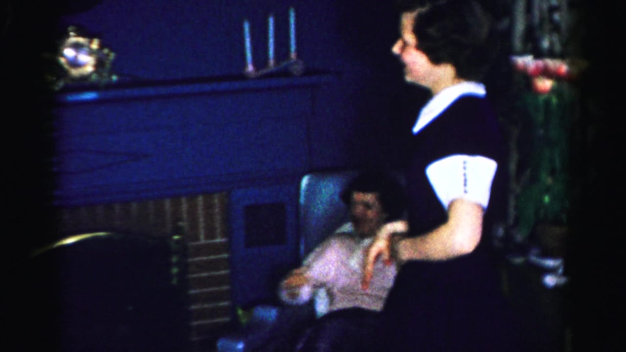 A woman in a dark dress with white collar pivots in a dimly lit mid-1950s domestic interior; a seated woman is visible in the background beside dark wood furniture.