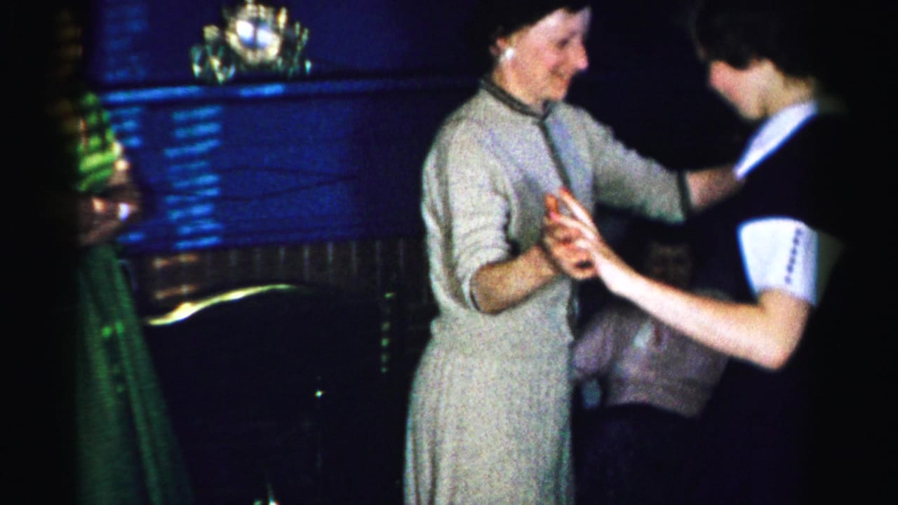 Two women in mid-1950s dress hold hands and dance in a domestic interior; the woman in a gray dress smiles broadly as a green drape and dark wood furniture fill the background.