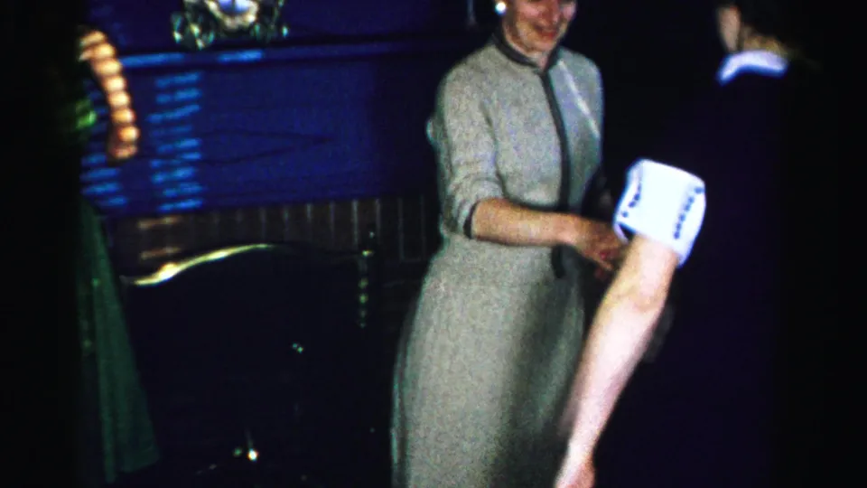 A woman in a gray 1950s dress laughs as she dances with another woman in a dim domestic interior; a warm orange lamp illuminates the left edge of the frame.