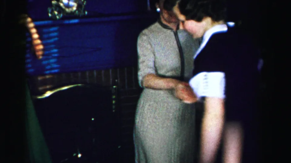 Two women in 1950s attire dance together in a private domestic room; the woman in a dark dress with white cuffs looks down as warm and cool light mix across the frame.