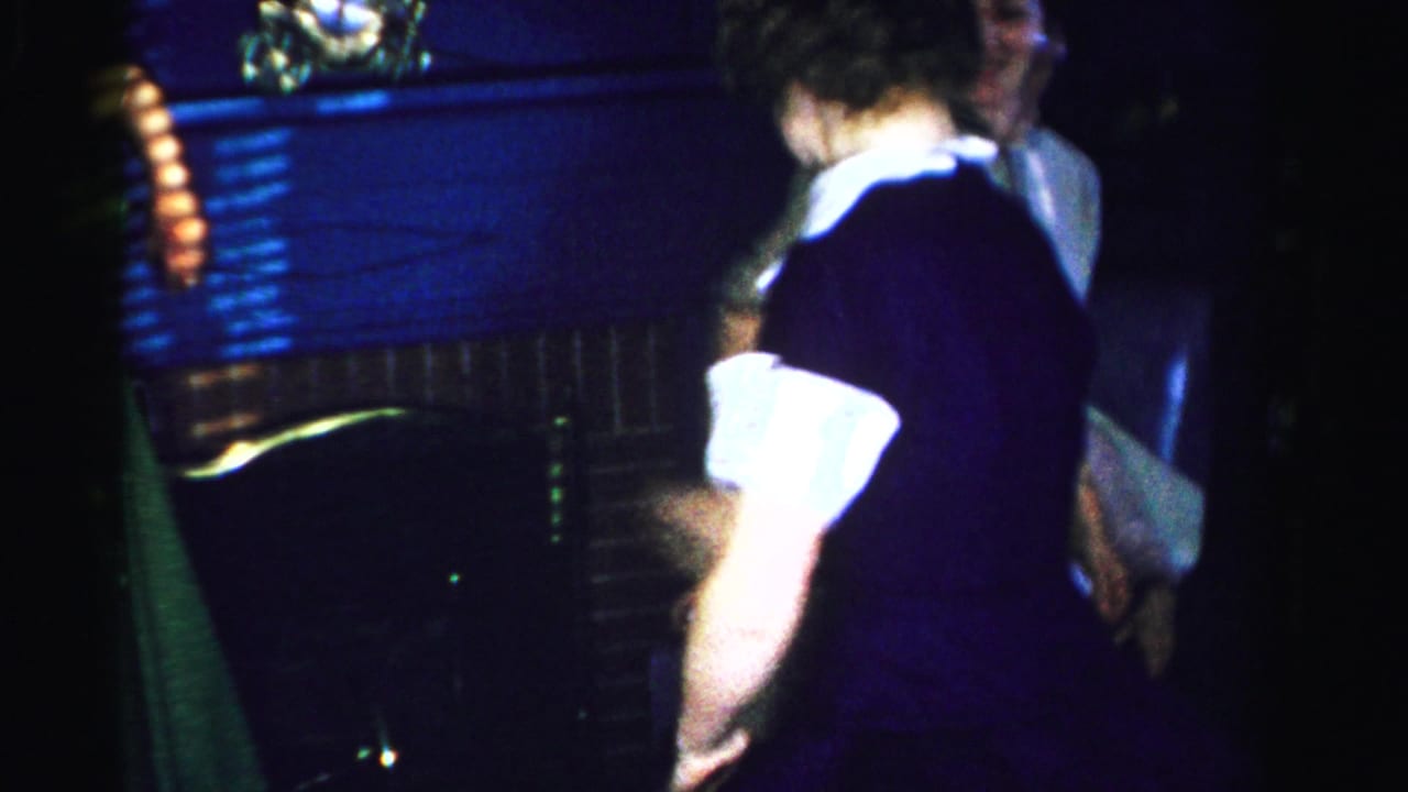 Woman in dark dress with white collar dancing, back to camera, brick fireplace mantel in background
