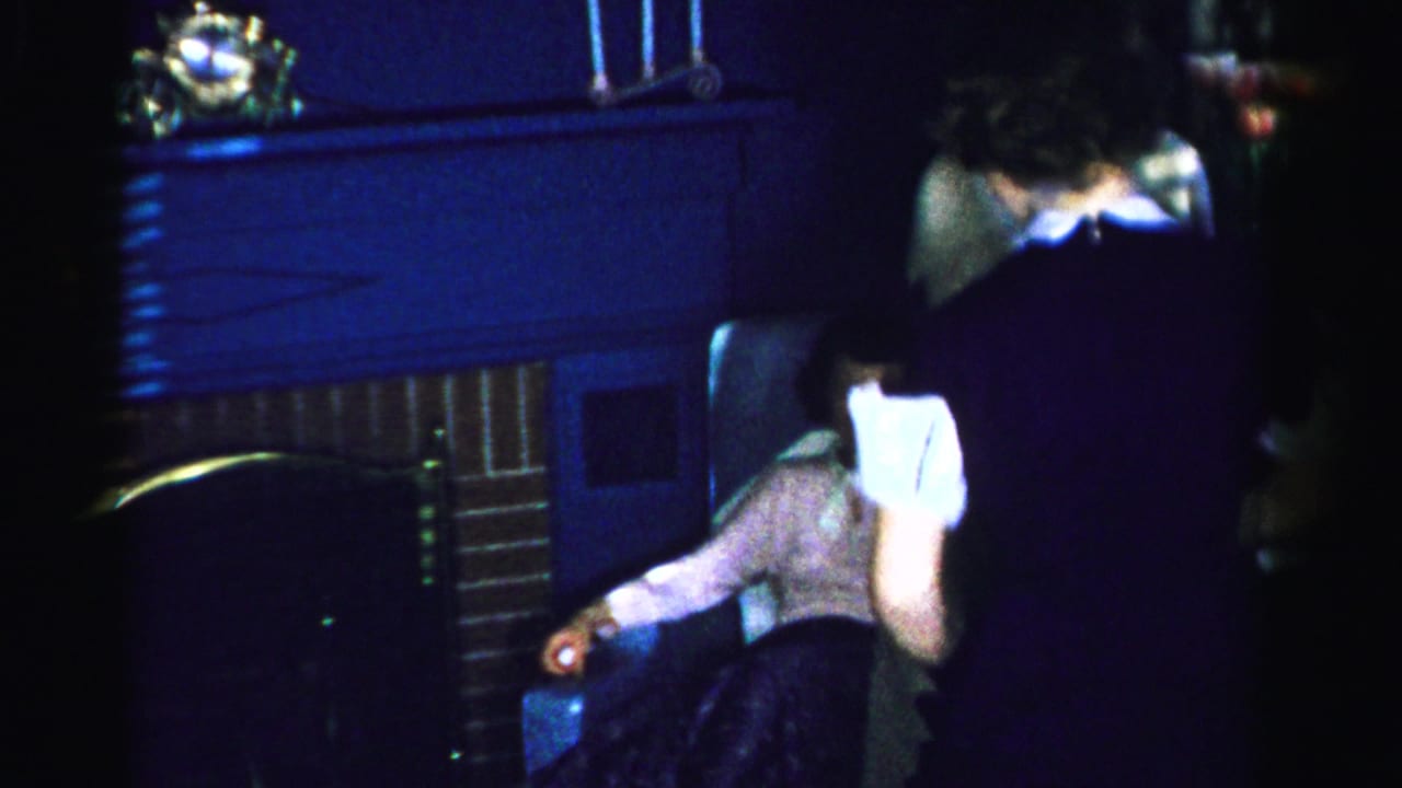 Two women at a mid-1950s indoor gathering, one in dark dress with white collar leaning forward, another in lighter clothing; brick fireplace mantel visible