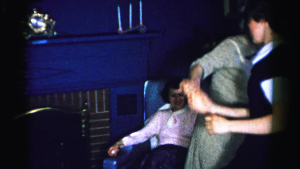 Two women dancing or interacting at a mid-1950s home gathering, one in dark dress with white collar; third figure partially visible low in frame; brick fireplace mantel behind