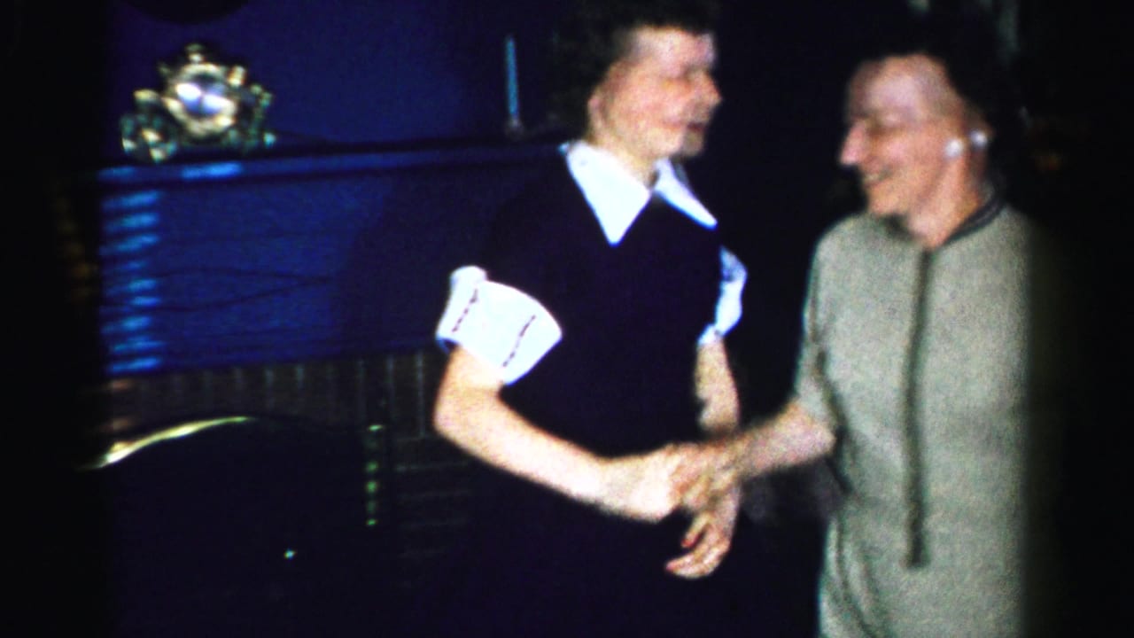 Two women clasp hands and smile at each other in a living room beside a fireplace mantel.