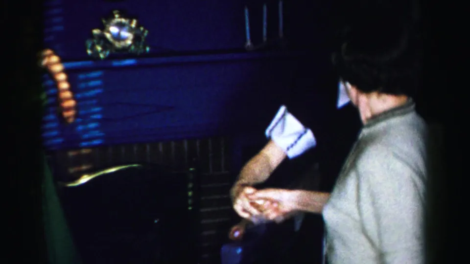 Close handheld shot of two women mid-dance; woman in dark dress with white collar and cuffs in foreground, another in a light dress behind; mid-1950s living room.