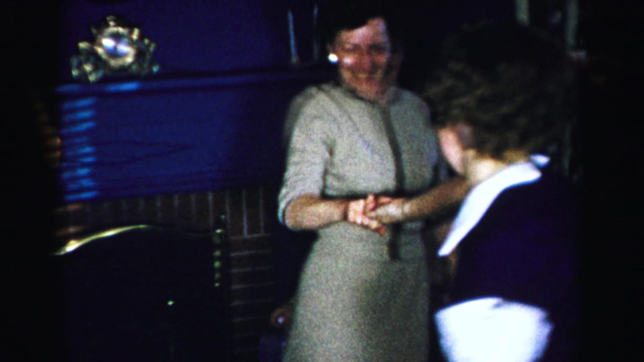 Two women face each other in a dimly lit room; one in a gray zip-front dress is smiling, the other in a white-collared outfit, appearing to dance or greet.