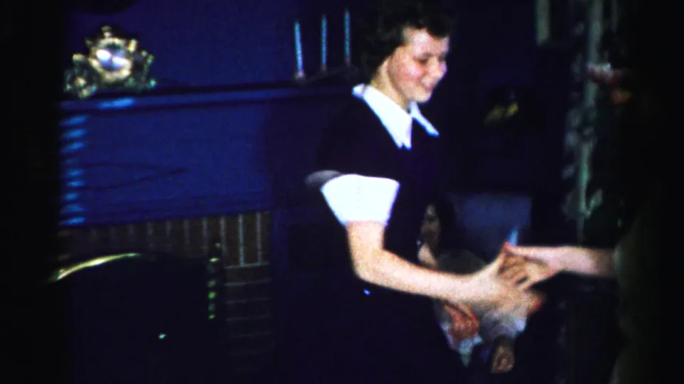 Close view of a smiling woman in a dark dress with white collar reaching out, likely mid-1950s Albany New York; additional figures are partially visible at the right frame edge.