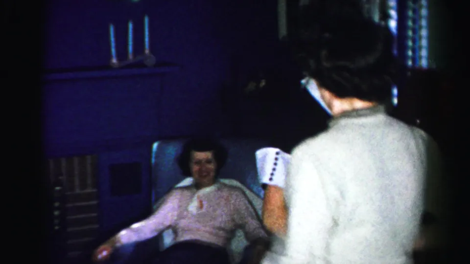View from behind two women at a mid-1950s indoor gathering; a third woman in a pink shirt seated in an armchair smiles, with candlesticks and dark wood furniture visible behind her.
