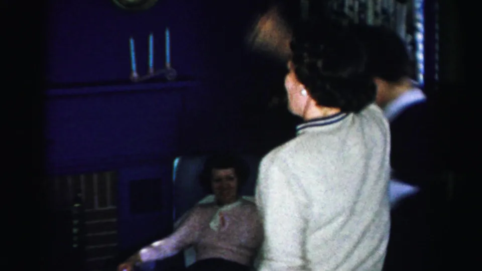 Indoor gathering circa mid-1950s Albany New York; a woman in a pink shirt seated in an armchair smiles as two others stand in the foreground with candlesticks visible behind her.