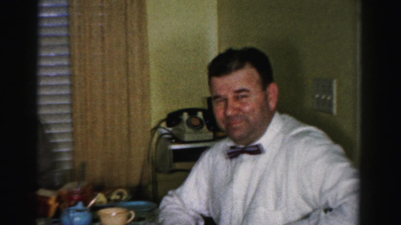 Man in white shirt and bow tie smiling candidly during a kitchen lunch gathering, circa mid-1950s; rotary telephone visible on surface behind him.