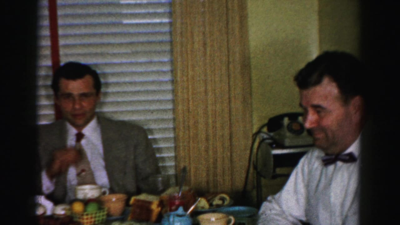 Two men at kitchen table; suited man with tie gestures near the table; colorful mugs and wicker basket visible; rotary telephone in background.