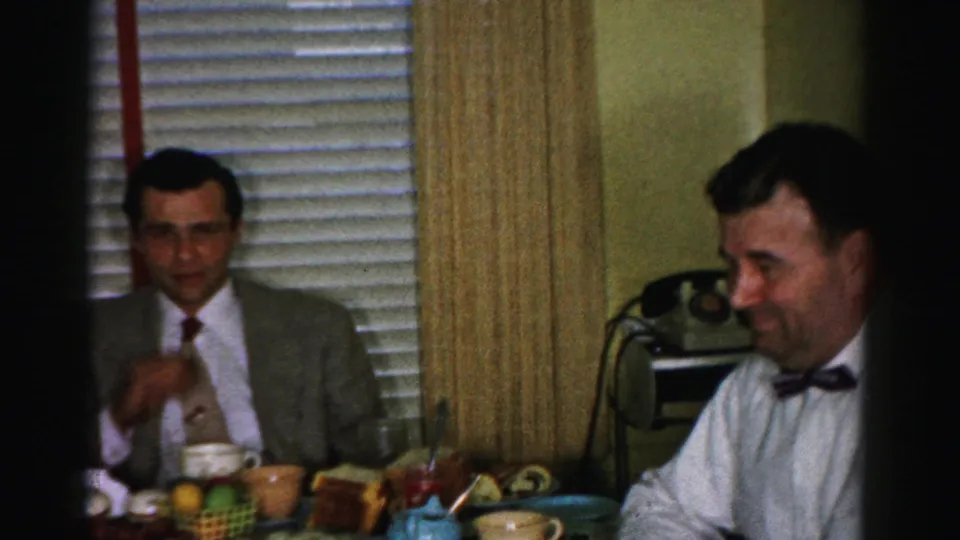 Two men at a kitchen table during a lunch gathering, circa mid-1950s; suited man gestures while bow-tie man is partially visible at left edge.