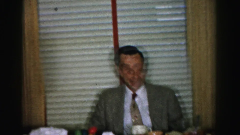 Suited man seated at kitchen table with a subdued expression during a mid-1950s domestic lunch gathering; colorful cups visible on table.