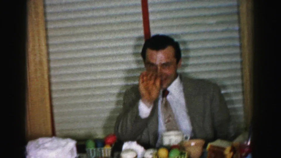 Suited man covers his face in laughter at a kitchen table during a mid-1950s domestic gathering; colorful table setting visible in foreground.