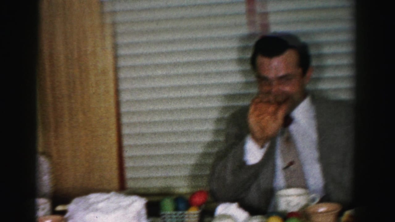 Man in gray suit covers face while laughing at a 1950s Easter table with colorful dyed eggs, ceramic cups, and venetian blinds behind him, Albany, New York area.