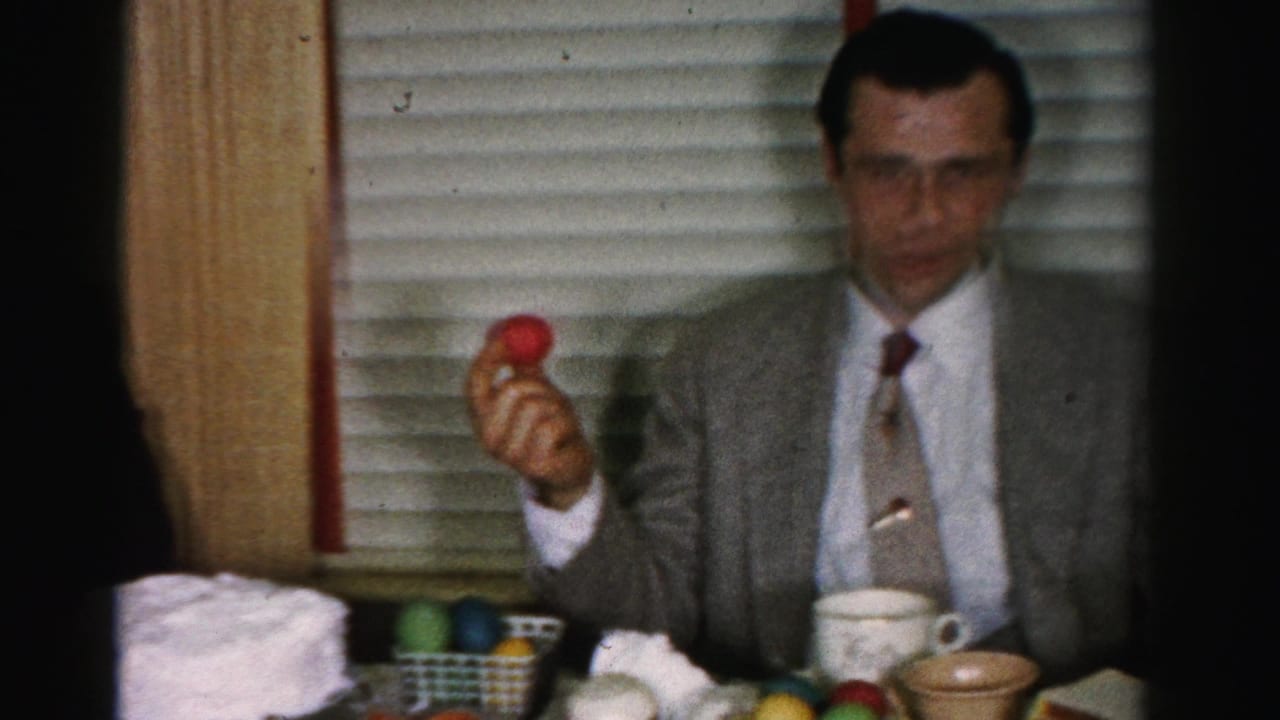 Man in gray suit and glasses holding a red Easter egg aloft at a table with a basket of colored eggs and ceramic cups.