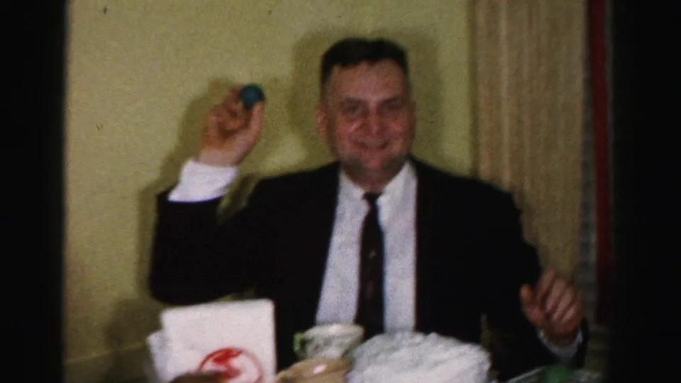 A second man in a dark suit holds a blue dyed egg at the Easter table; a logo-marked napkin holder is visible in the foreground, 1950s setting.