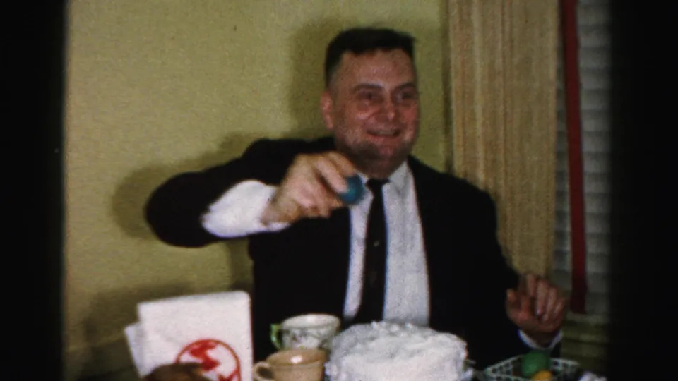 Man in dark suit gestures toward camera with a dyed Easter egg; a white frosted cake and logo-marked napkin holder are visible on the table, 1950s Easter celebration.