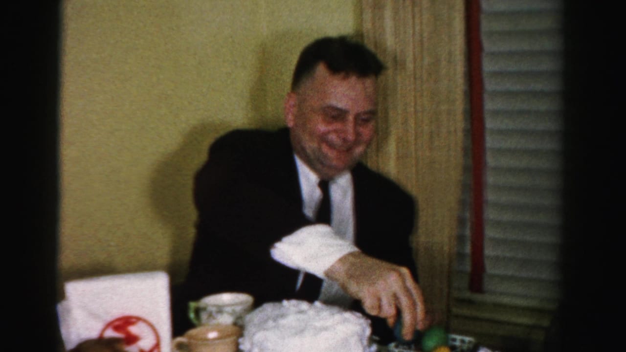 Middle-aged man in dark suit and tie at a set table, smiling downward at colored objects and tableware, venetian blinds behind him.