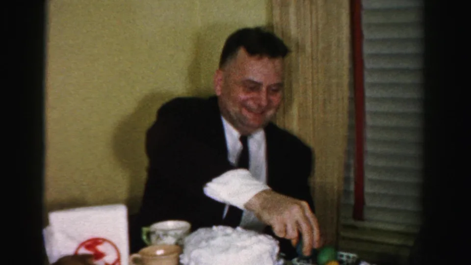Middle-aged man in a dark business suit and narrow tie at a dining table in what appears to be a mid-1950s Albany, New York interior, smiling at items on the table.