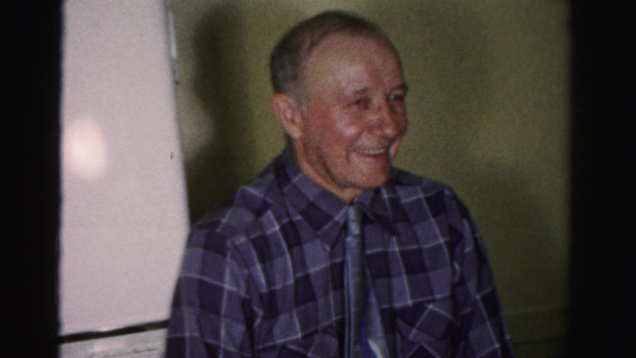 Older man in a plaid sport shirt and tie smiling warmly in a domestic interior, consistent with mid-1950s Albany, New York home-movie footage.