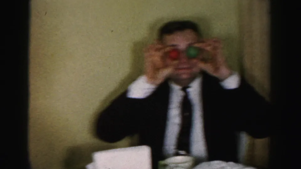 Man in a dark business suit and tie performing a ball-over-eyes table gag, mid-1950s 8mm home-movie footage consistent with Albany, New York setting.