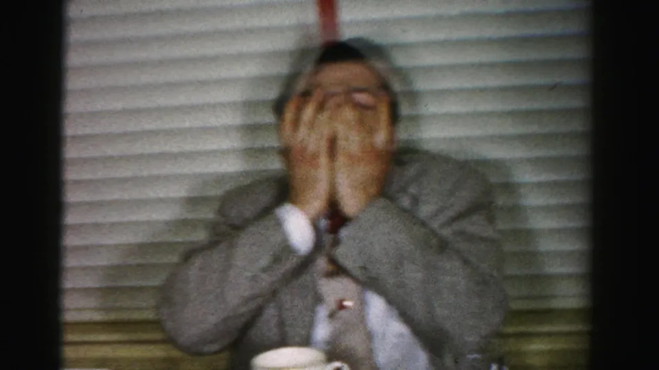Motion-blurred frame of a man in a gray suit covering his face with both hands, mid-1950s 8mm home movie consistent with Albany, New York interior.