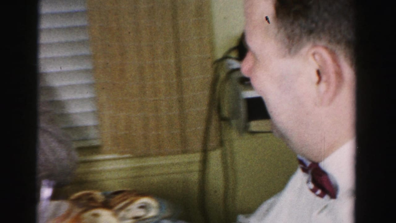 Profile close-up of a man in white dress shirt and dark tie; pastries and wall-mounted telephone visible in background.