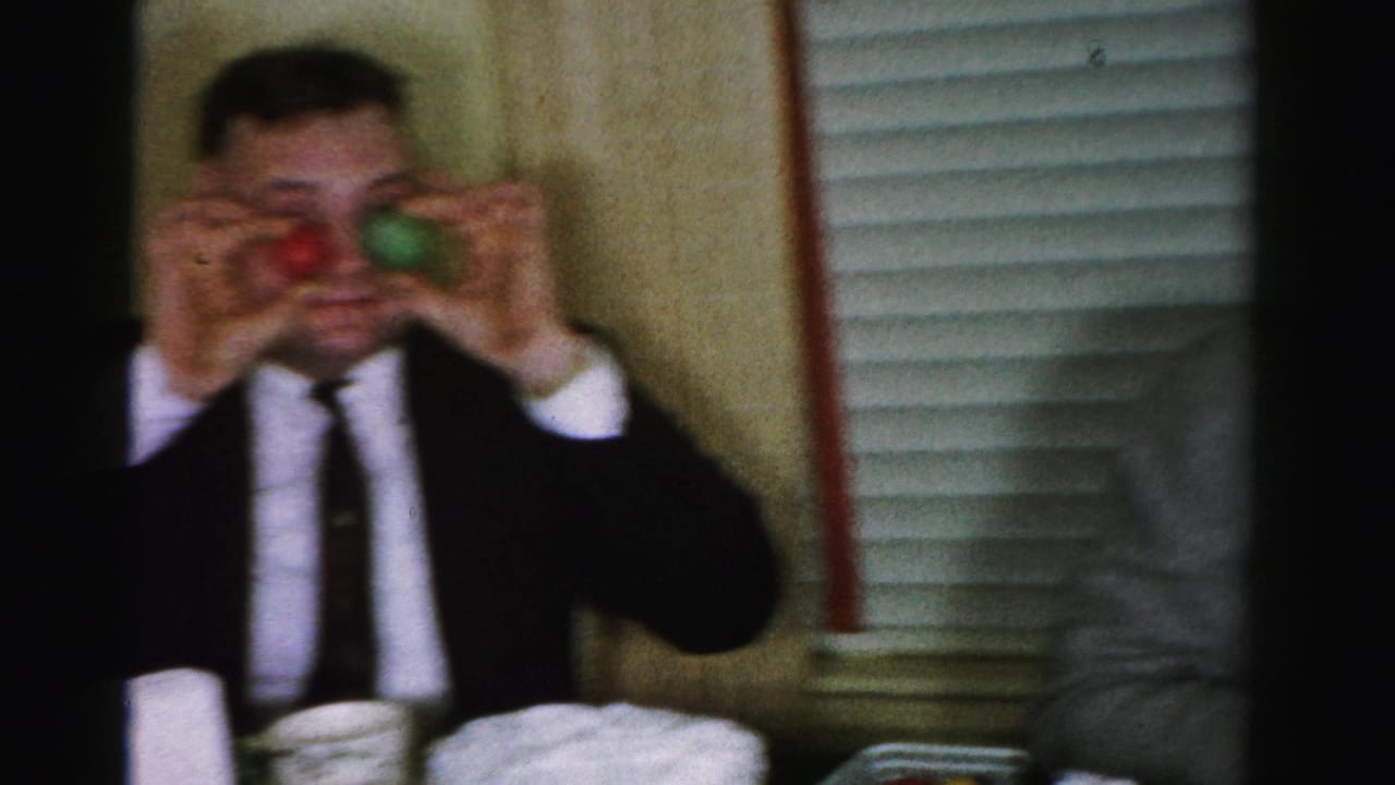 Man in dark suit and narrow tie performing a comic gesture with small colored objects at a mid-1950s birthday party; white layer cake and window blinds in background, Albany, NY, c. 1956.