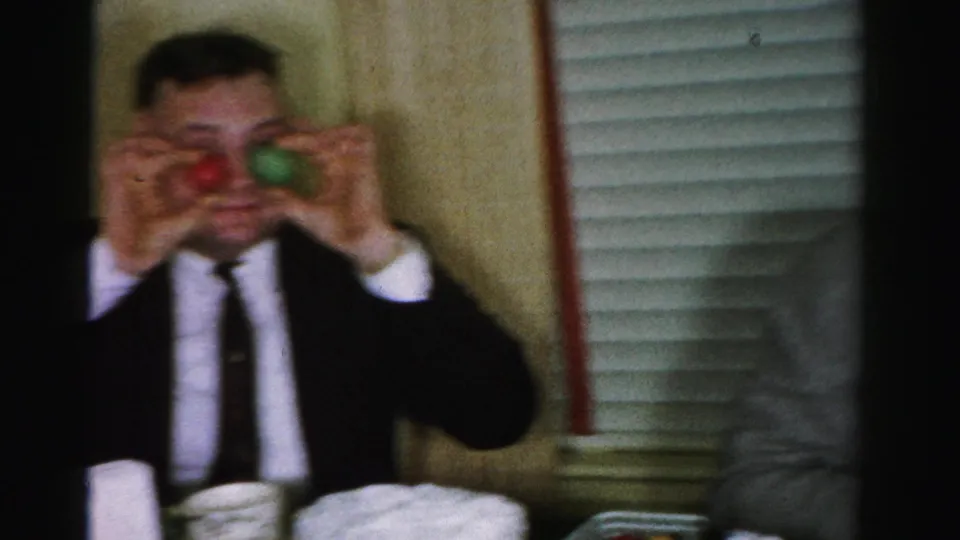 Man in dark suit and narrow tie performing a comic gesture with small colored objects at a mid-1950s birthday party; white layer cake and window blinds in background, Albany, NY, c. 1956.