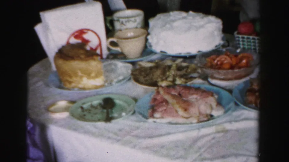 Close shot of a mid-1950s birthday party table with white-frosted layer cake, decorated pastry with red motif, sliced cold cuts, fruit, and cups; Albany, New York, c. 1956.