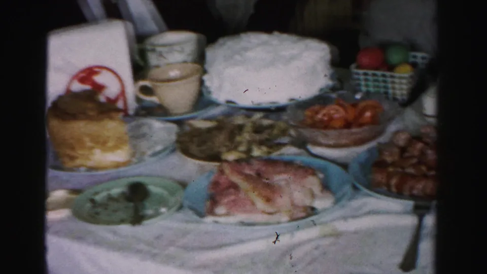 Mid-1950s Easter holiday table in Albany, New York, showing a white frosted cake, sliced ham, sausages, and dyed eggs in a checkered basket on a white linen cloth